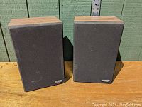 Front view of pair of speakers with black fabric grilles on wooden surface and green background, showing condition of front panel and grille.