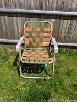 Front view of vintage lawn chair showing woven green and beige webbing seating and curved aluminum arms.