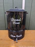 Front view of the large black speckled enamelware clam steamer pot with white text labels 'Steamed Clams' on upper pot and 'Clam Broth' on base, showing the spigot