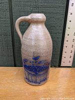 Front view of tan stoneware bottle with handle and cobalt blue blueberry basket design alongside ruler showing height approximately 10 inches.