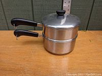 Side view of stacked two quart saucepan and steamer pan with lid on top showing black handles and stainless steel body.