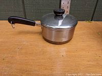 Side view of the vintage Revere Ware saucepan with lid placed on a wooden surface outdoors next to a ruler for scale.