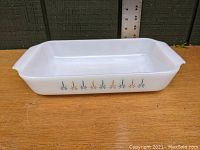 Rectangular white opaque glass baking dish with decorative flower pattern on side, placed on a wooden surface against a green wall with measuring scale behind it.