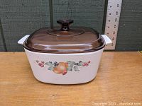 Photo of white ceramic casserole with fruit and berry pattern on front and brown Pyrex glass lid on top.
