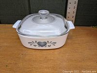 Front view of the white square CorningWare casserole with blue heart pattern, glass lid in place, placed on wooden surface with ruler in background.