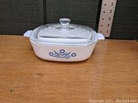Side view of white CorningWare casserole dish with blue cornflower design and glass lid, placed on wooden surface with measuring stick behind.