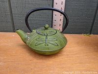 Green cast iron Japanese tea kettle with dragonfly motifs, side view showing spout, handle, and lid.