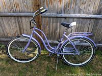 Side view of lavender Schwinn Huntington women's cruiser bicycle with whitewall tires, black and white saddle, rear rack, and kickstand.