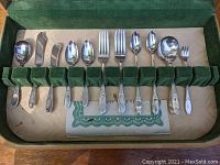 Close-up of bottom row of flatware showing butter spreaders, sugar spoon, teaspoons, dinner forks, soup spoons, and cocktail forks in cloth-lined slots with detailed handles.