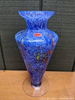 Blue mouth-blown Murano art glass vase with wide neck, flared rim, and conical base resting on a clear footed base. Visible color inclusions of yellow and orange in deep blue speckled background.