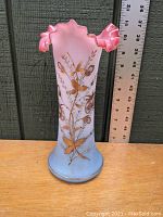 Photo of the entire Victorian blown satin glass vase showing pink to blue gradient and gilded foliate design with ruffled rim, next to ruler for size reference.
