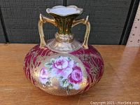 Front view of Victorian porcelain vase with two handles, burgundy body, heavy gilding, and hand-painted pink roses in oval cartouche.