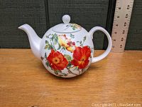 Front and side view of white bone china teapot decorated with large red and yellow poppy flower designs on wooden surface with a ruler in background.