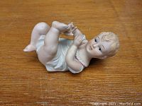 Close-up front and side view of Victorian-style bisque porcelain baby figurine lying on back holding right foot, with visible facial features and garment detail.