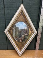 Full view of diamond shaped white and gold frame with classical ruins print inside, placed next to a ruler for scale.