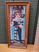 Full view of framed vintage mid-century big eyes harlequin girl print in masquerade costume standing against a wooden background with measuring ruler for scale.