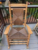 Front view showing wooden porch rocker with woven rush seat and backrest, on outdoor deck area.