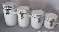 Four white ceramic canisters of varying sizes with metal clip lids and side handles placed side by side on a gray surface.