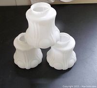 Three camphor glass light shades arranged with one standing on top and two on the bottom showing the shape and opaqueness against a dark background.