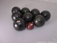 Eight black bocce balls and one reddish-brown pallino ball arranged on a gray surface showing the white inlays on balls