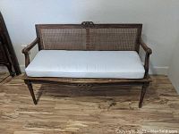 Full view of wooden settee bench with cane back and white seat cushion on wood flooring.