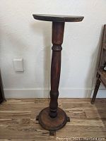 Full view of the walnut Victorian plant stand showing the round top, single turned central pedestal, and round base with feet on wooden floor and white textured wall background.