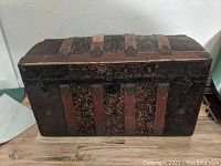 Front view of the oak slat domed top trunk showing the domed top, leather covering with metal reinforcements, and two clasps on the front.