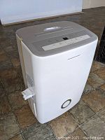 White Frigidaire dehumidifier on tiled floor with a side drainage hose visible.