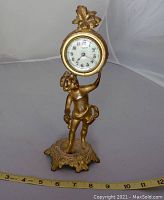 Front angle view of antique gilt cherub desk clock showing decorative base, cherub figure, and clock face with white dial and black numbers.