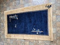 Full view of rectangular cobalt blue and beige Chinese silk rug with floral and bamboo design on stone floor.