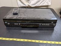 Front view of the Toshiba black DVD and VCR combo player unit with remote control placed on top, showing the front panel and control buttons.