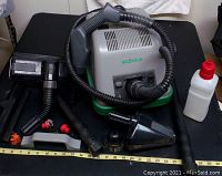 Photo showing SGT Steam Cleaner main unit with attached hose and various black plastic nozzle attachments plus white plastic bottle with red cap on black surface with measuring tape.