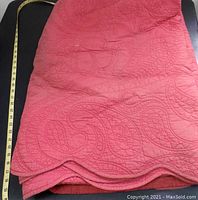 Close-up image showing the folded red quilted blanket with visible intricate quilted patterns and a measuring tape indicating size.