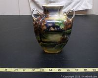 Front and side views of Noritake vase showing the scenic hand painted design with small house, water, trees, and blue sky. Two upright double handles on each side. Gold trim detail on rim and foot.
