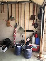 Photo showing various hanging gardening tools including metal and plastic leaf rakes, brooms, garden hoe, shovels, and a flag on pole
