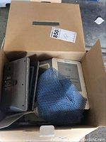 Box of items including a weather station, Amazon Kindle e-reader, and a grey mesh sleeve.