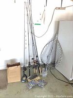 Seven fishing poles with reels, two fishing nets, fillet knife, and Banjo Minnow lure packages on the floor against a garage wall.