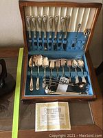 Wooden chest open showing organized silver plated knives, forks, spoons, and serving utensils with blue fabric lining.