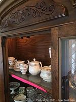 China and glassware displayed in a wooden cabinet shelf, showing floral patterned white porcelain pieces, including teapot, creamer and sugar bowl.