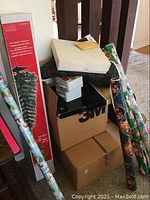 Various stacked boxes including a boxed artificial Christmas tree and rolls of Christmas wrapping paper with holiday prints.