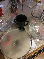 Photo showing assorted glassware including clear pedestal cake plate, black handled glass pitcher, various clear glass vases and bowls on a surface, tagged with lot numbers.