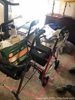 Red walker with seat and backrest, along with black walker and package of adult diapers nearby. Carpeted floor with pink tape markings.