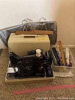Vintage black Singer sewing machine sitting in the beige carrying case with hangers and garment bag behind it and a bin of sewing accessories to the side on a carpeted floor.