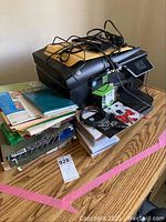 Front and top view of black HP Officejet 6600 printer with cables on top and stacks of papers, envelopes and folders underneath on wooden surface.