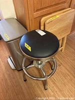 View of the trash can, garage stool with black leather seat, and two wood TV trays stacked against wall.