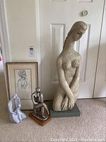 All three statues and the framed pencil sketch of a nude woman by Alfred Czerepak placed on carpeted floor against a white door.