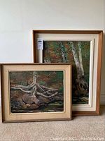 Two framed acrylic paintings leaning against a wall, smaller foreground painting shows exposed tree roots with rocks and water.