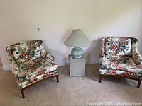 Pair of floral fabric upholstered chairs shown side by side with a lamp in between, displaying overall style and condition.