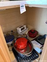 Image of cabinet corner showing a red enamel pot with lid, roasting pans, glass containers, and other kitchen items stacked.