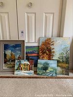 Five nature scene paintings including framed and unframed, varied sizes, placed against closet doors on carpeted floor.